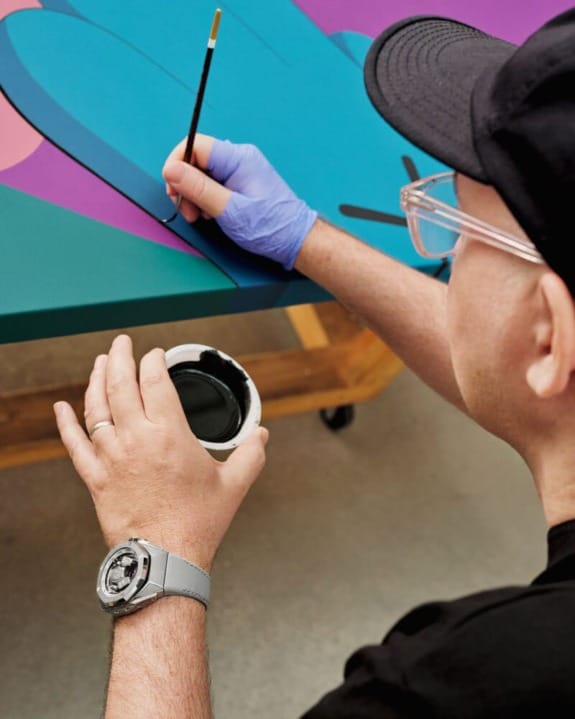 Artist painting vibrant abstract piece on canvas with brush in hand, wearing glasses, cap, and smartwatch.