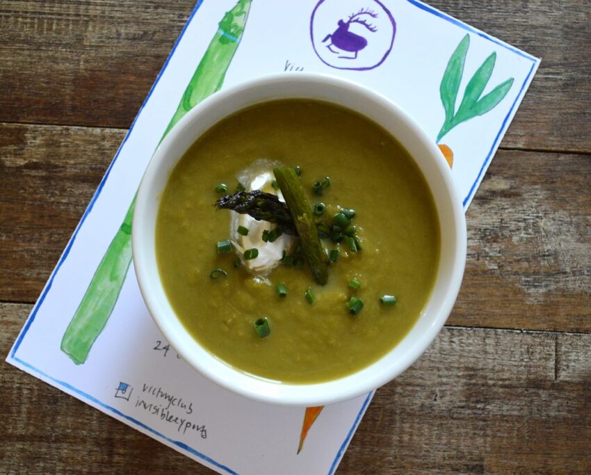 Bowl of green soup garnished with asparagus and chives, on a wooden table with a colorful vegetable illustration underneath.