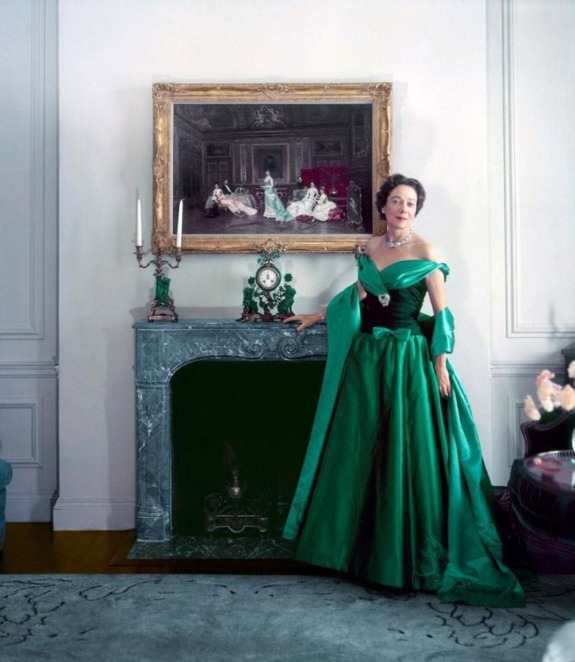 Woman in elegant green gown stands by a fireplace adorned with a vintage painting and decorative clock.
