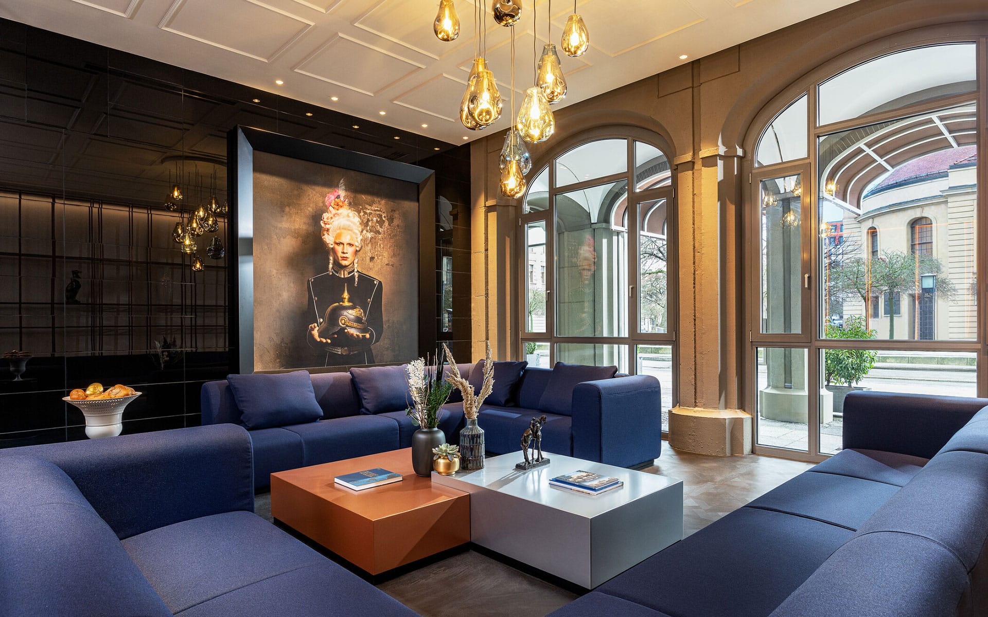 Modern lounge with navy sofas, contemporary artwork, decorative lights, and large windows overlooking the building exterior.