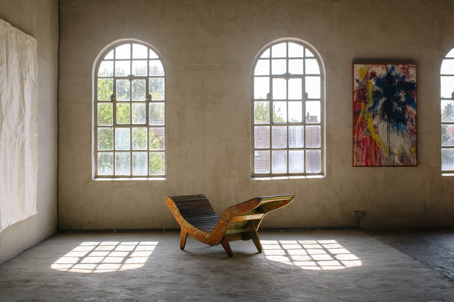 Sunlit room with three arched windows, a wooden chaise lounge, and a colorful abstract painting on the wall.