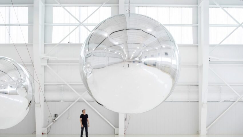 Person standing next to a large reflective silver sphere in a bright, industrial space.