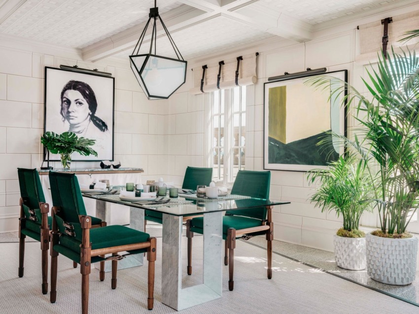 Modern dining room with green chairs, glass table, large paintings, geometric pendant light, and potted plants near windows.
