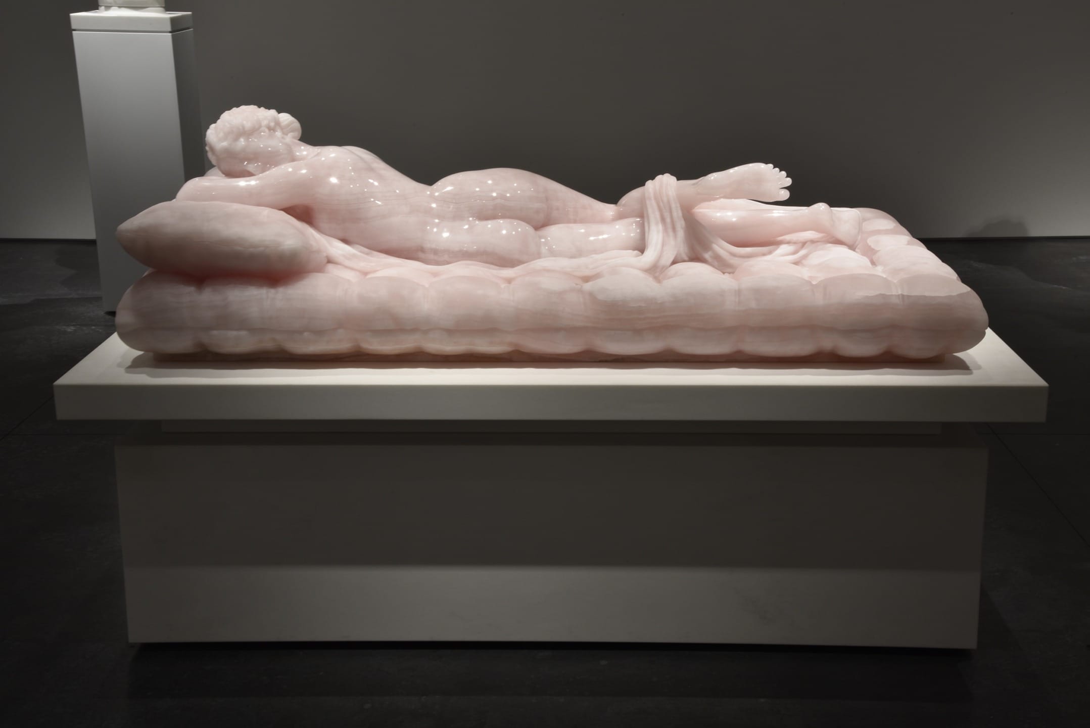 Reclining marble sculpture of a human figure on a pillow, displayed in a dimly lit gallery setting.