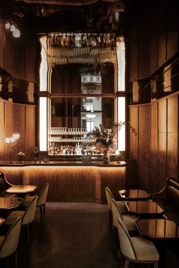Elegant, dimly lit bar with reflective ceiling, sleek wooden furniture, and a stocked backlit bar counter with floral arrangement.