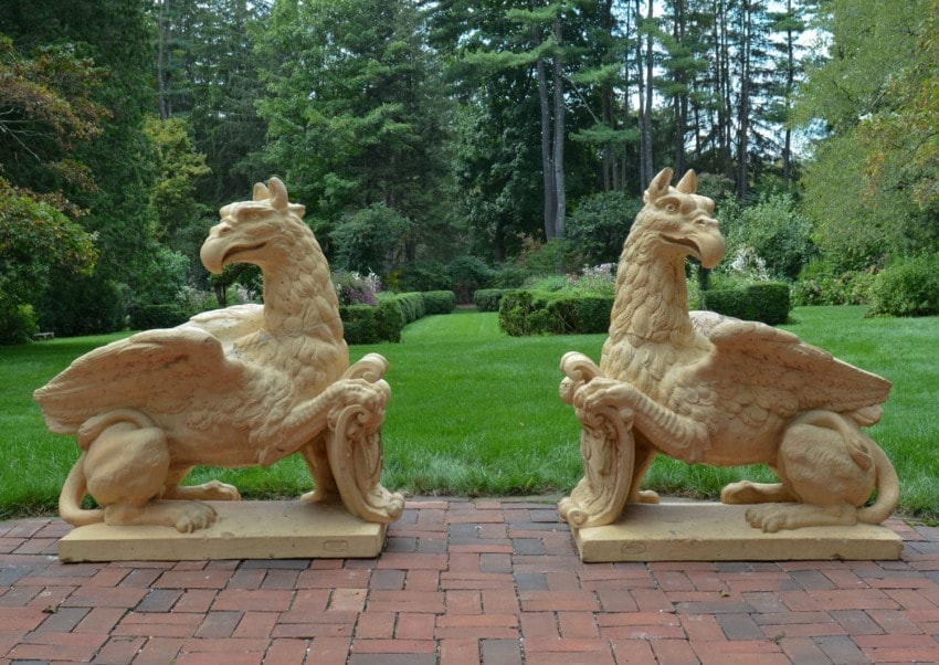Two stone griffin statues on a brick path in a lush garden setting with tall trees in the background.
