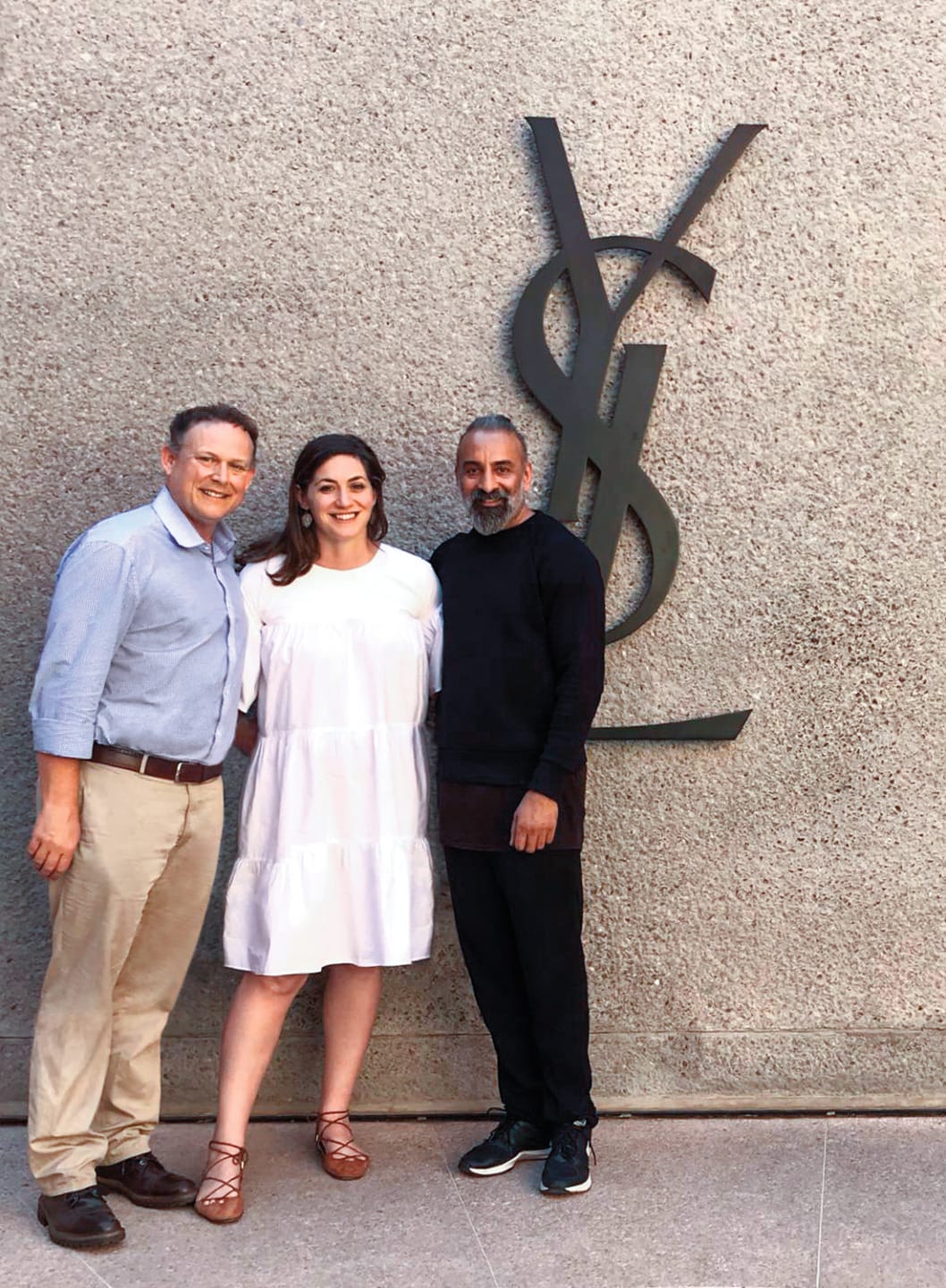 Three people posing in front of a large YSL logo on a textured wall.