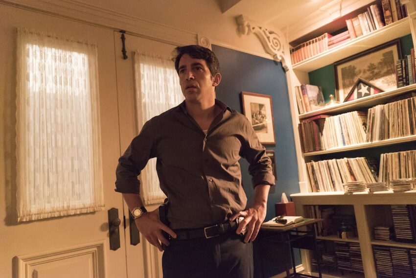 Man standing in a room with records and books, hands on hips, wearing a brown shirt, near a door and well-lit interior.