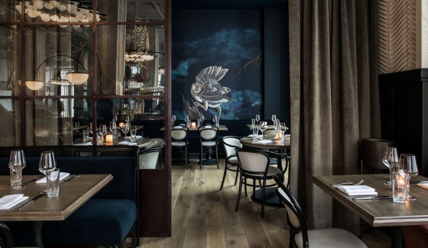 Elegant restaurant interior with wooden tables, chairs, dim lighting, and a mural of an owl on a dark blue wall.