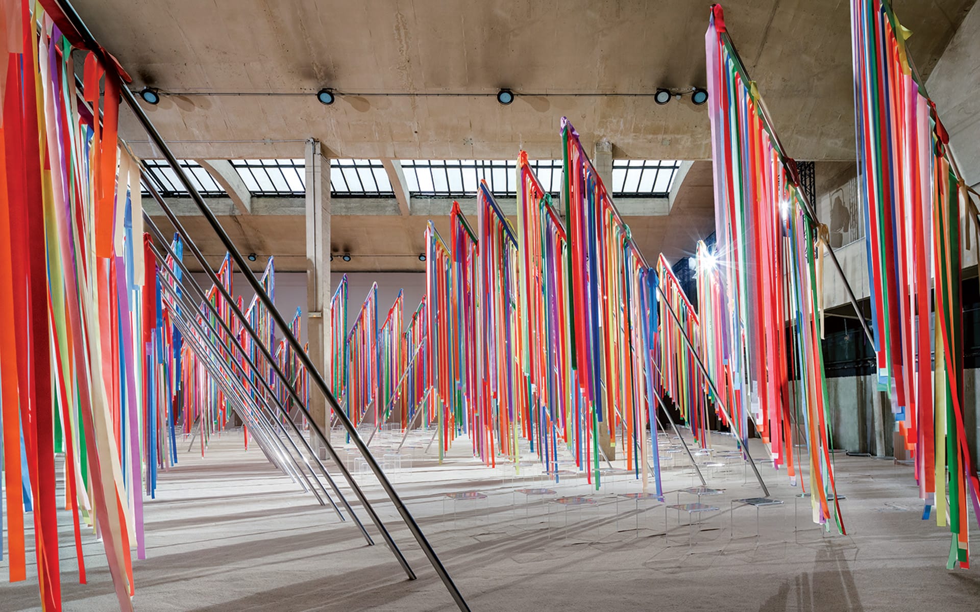 Colorful art installation featuring long vertical ribbons hanging from poles in an industrial-style gallery space