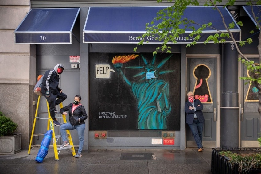 Three people standing by a mural of the Statue of Liberty holding a torch with "Help" sign, outside a building with blue awning.