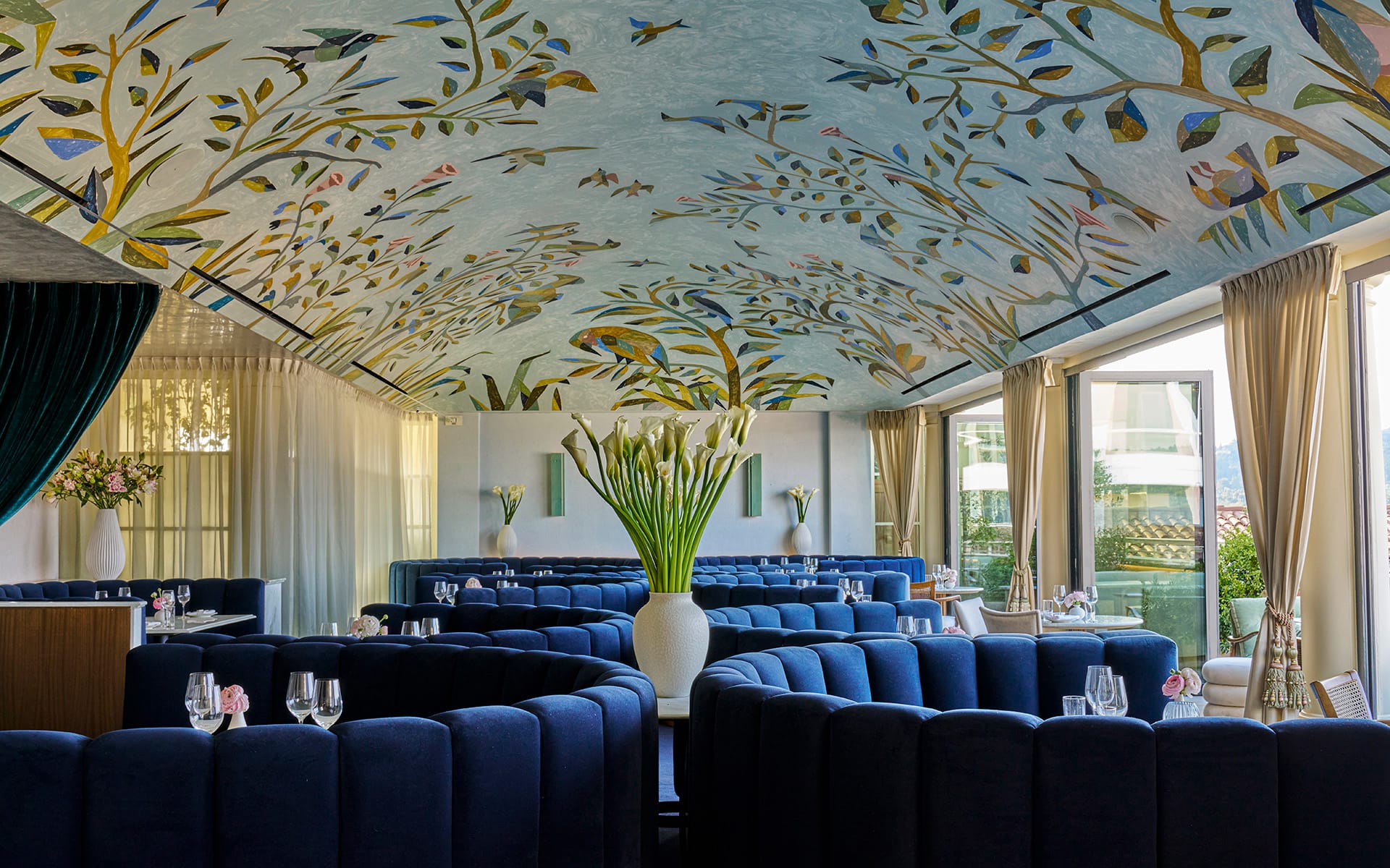 Elegant restaurant interior with blue velvet seating, artistic ceiling design, large vases and natural light from tall windows.