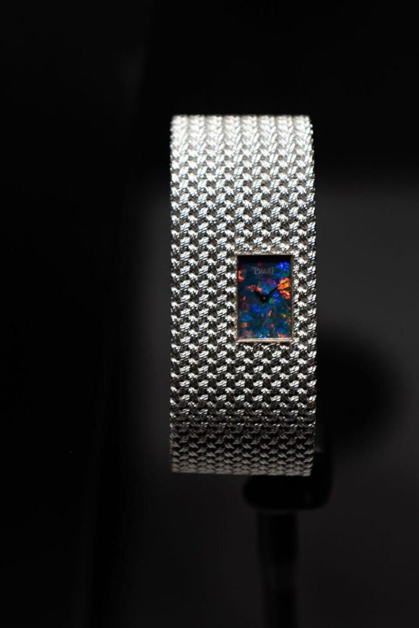 Silver mesh watch with a colorful opal face on a dark background.