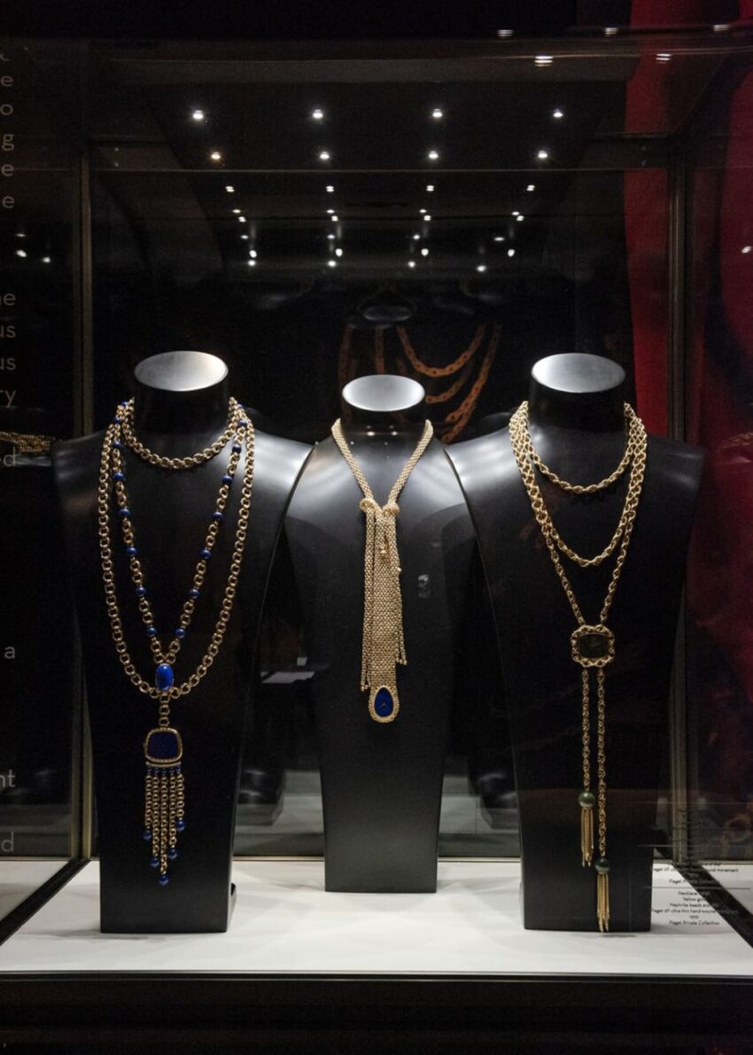 Three elegant necklaces on display with intricate designs and various gemstones on black stands in a well-lit glass case.