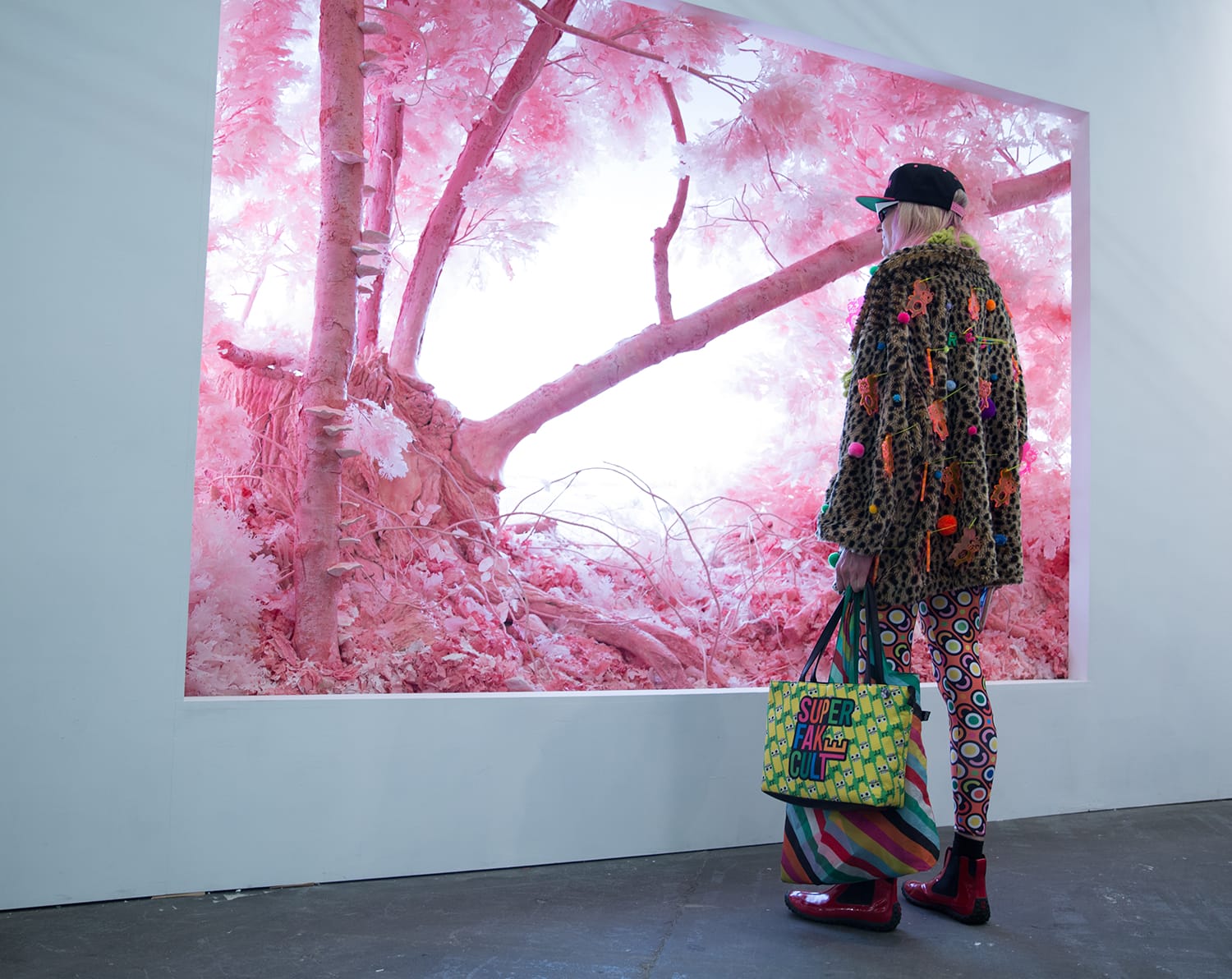 Person in colorful outfit admires large artwork of a vibrant pink forest scene on a gallery wall.