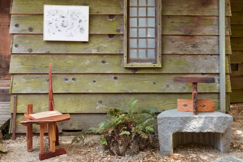 Wooden chairs and table set outside a rustic cabin with green wooden walls and a small plant at the base.