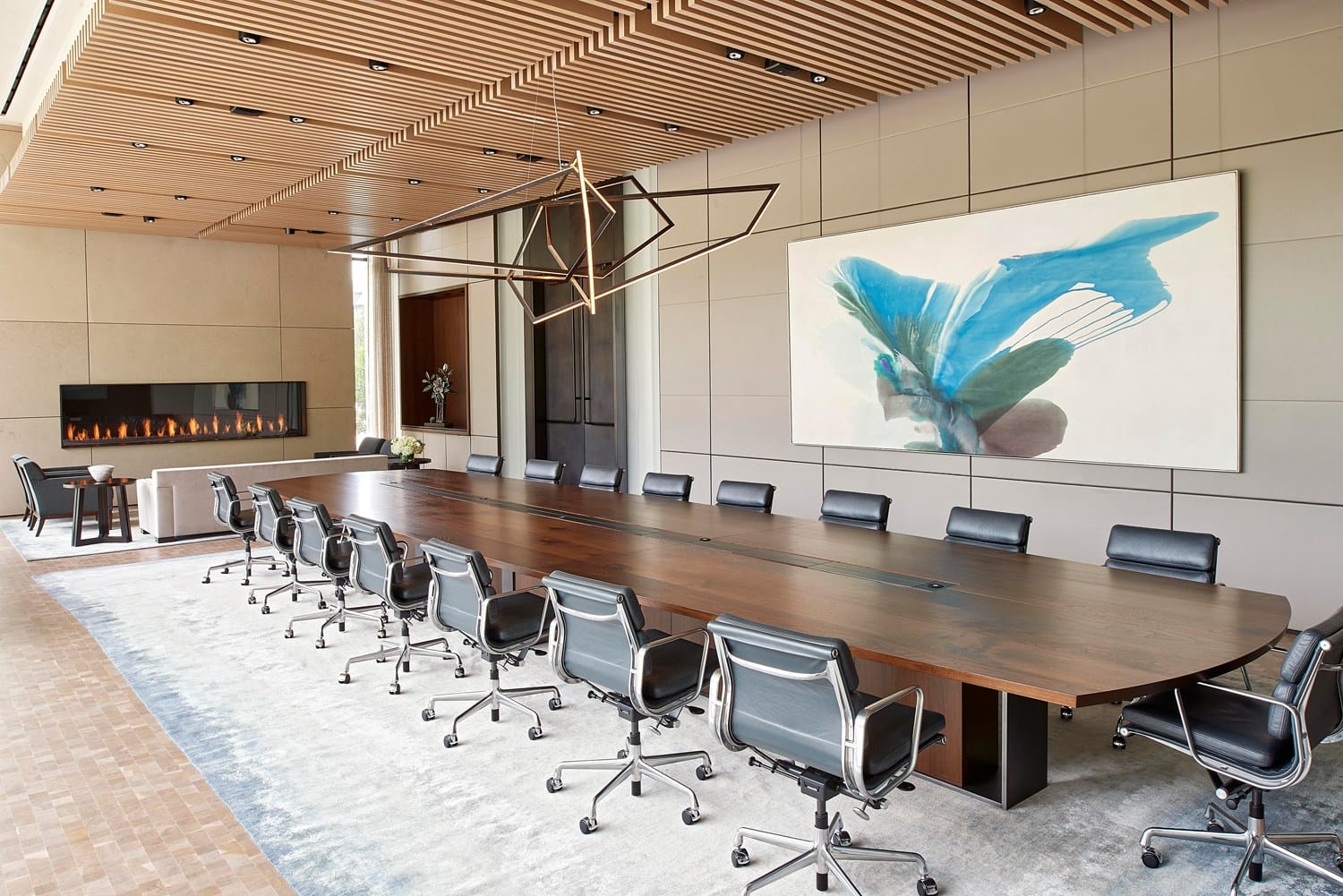 Modern conference room with a long wooden table, black chairs, geometric lighting, a large abstract painting, and a fireplace.