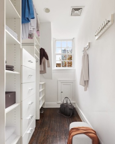 Compact white walk-in closet with shelves, hanging clothes, a window, and a bag on the floor near a small bench.