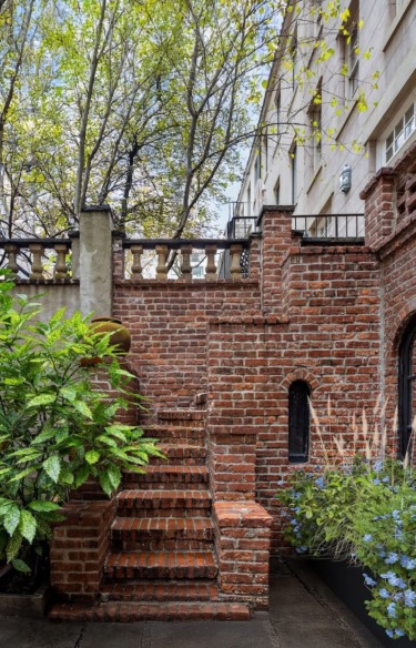 Brick staircase with lush green plants on an outdoor patio, surrounded by trees and an old building in the background.