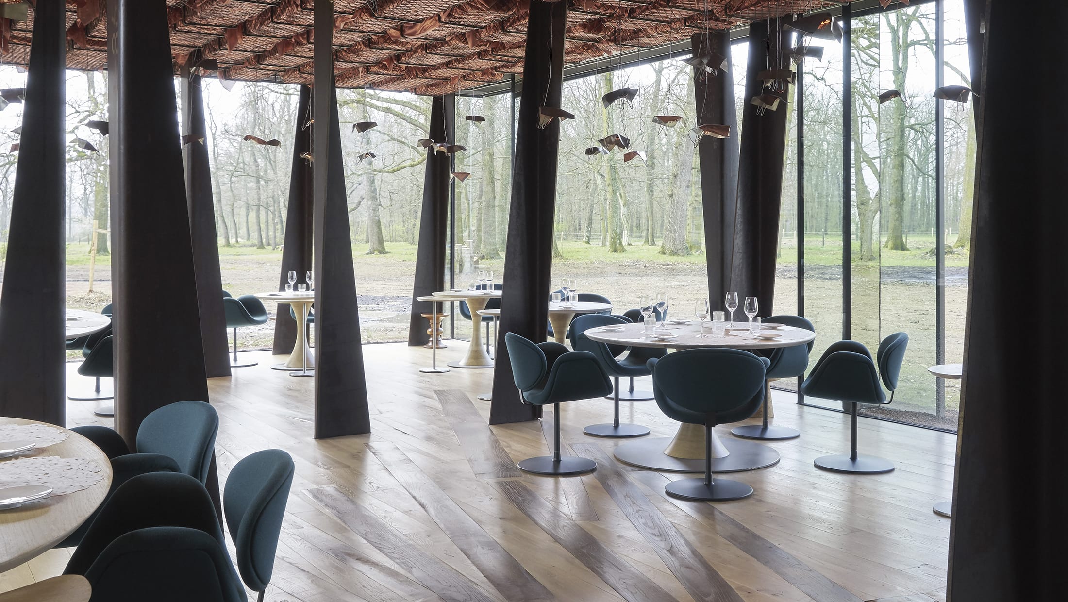 Modern restaurant interior with teal chairs, round tables, and large windows overlooking a wooded area.