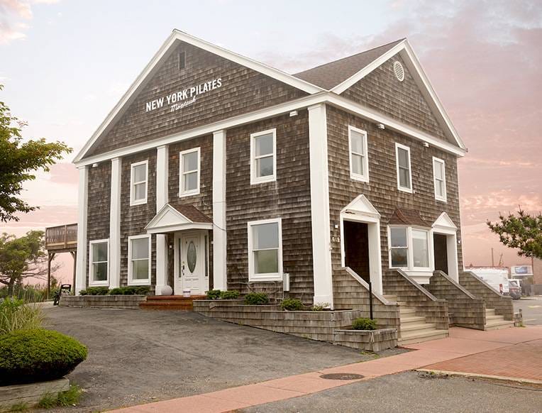 A rustic two-story building with a shingle facade and "New York Pilates" sign, set against a pink and blue sunrise sky.
