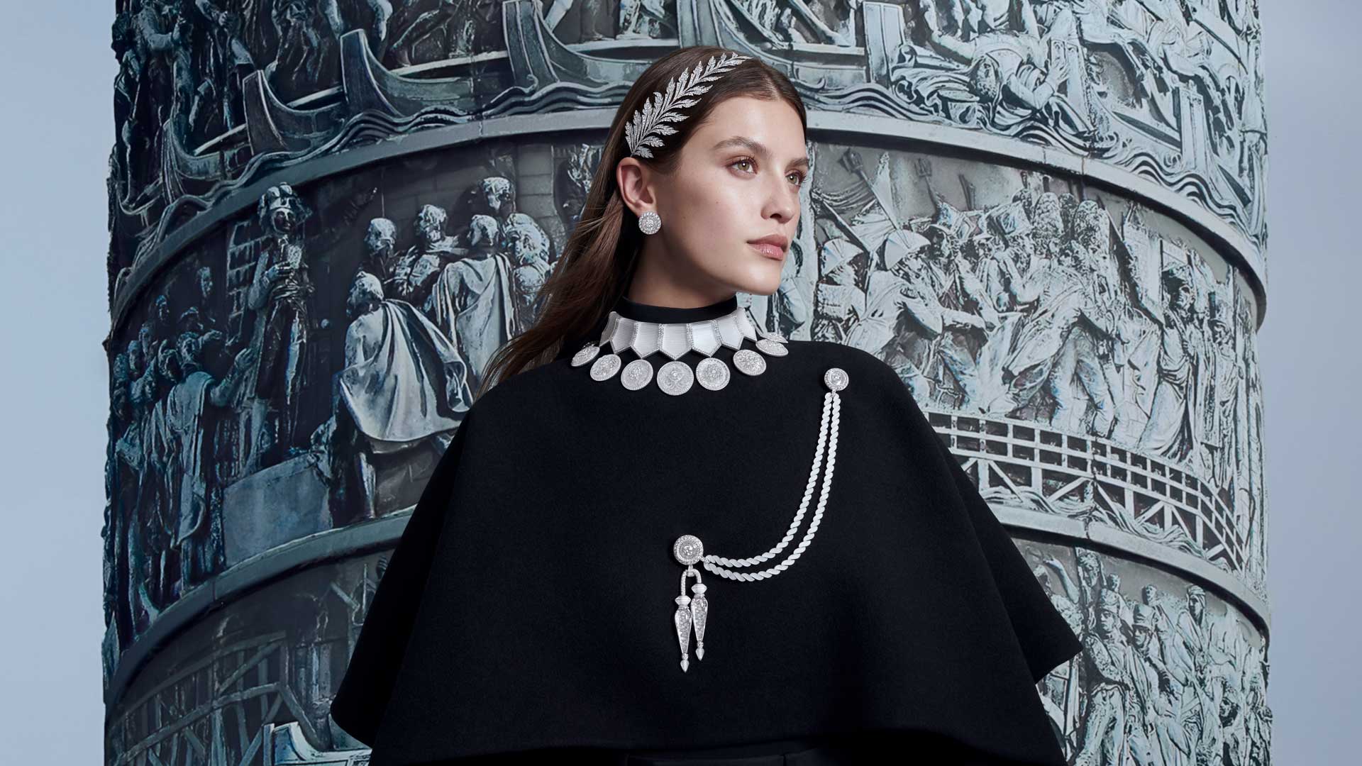 Woman in black cape with silver jewelry stands before a detailed sculptural wall, looking to the side.