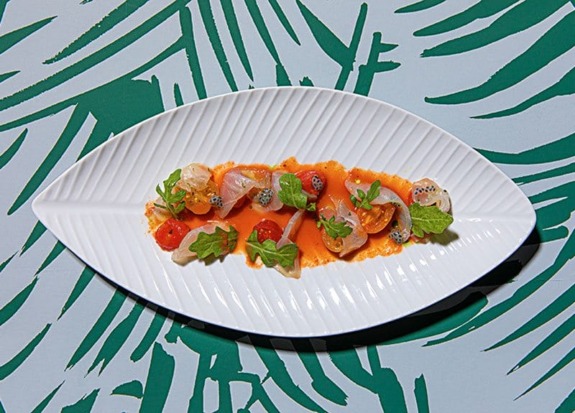 Plate of sliced fish with cherry tomatoes, arugula, and sauce on a leaf-patterned table.