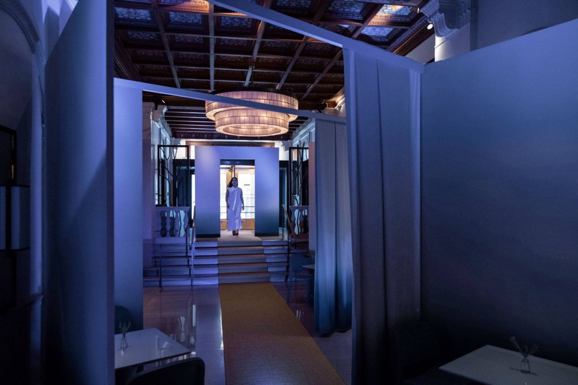 Dimly lit modern restaurant interior with a person in a robe standing near the entrance under a large circular chandelier.