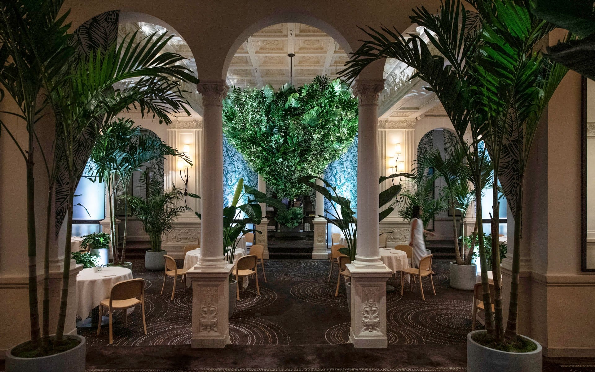 Luxurious dining area with elegant arches, lush greenery, and round tables set for guests in an ornate interior setting.