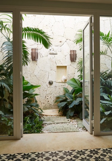 Outdoor shower surrounded by lush green plants and stone wall, viewed through open glass doors.