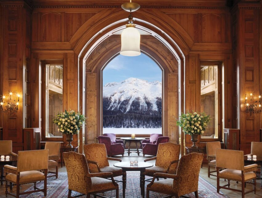 Luxurious wooden lounge with elegant chairs, large arched window view of snowy mountain landscape, floral arrangements and warm lighting.