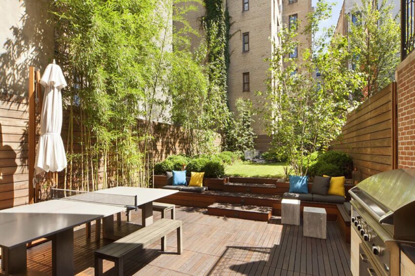 Outdoor urban patio with tables, benches, plants, and a barbecue grill under sunlight, surrounded by brick buildings.