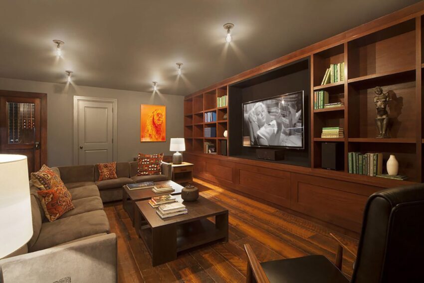Cozy living room with wooden floor, brown sectional sofa, wall-mounted TV, and bookshelves filled with books and decor.