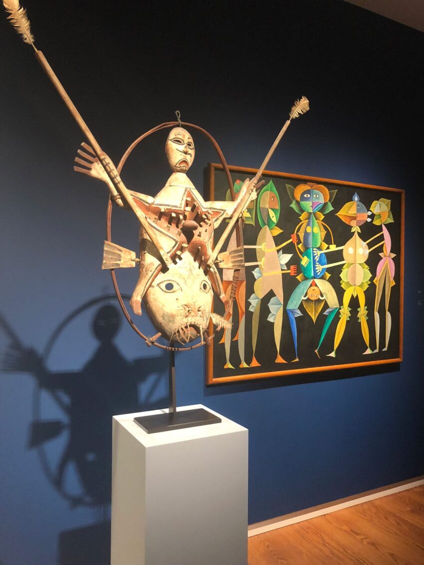 Exhibit of abstract wooden sculpture and colorful painting in a museum gallery with blue walls and wooden floor.