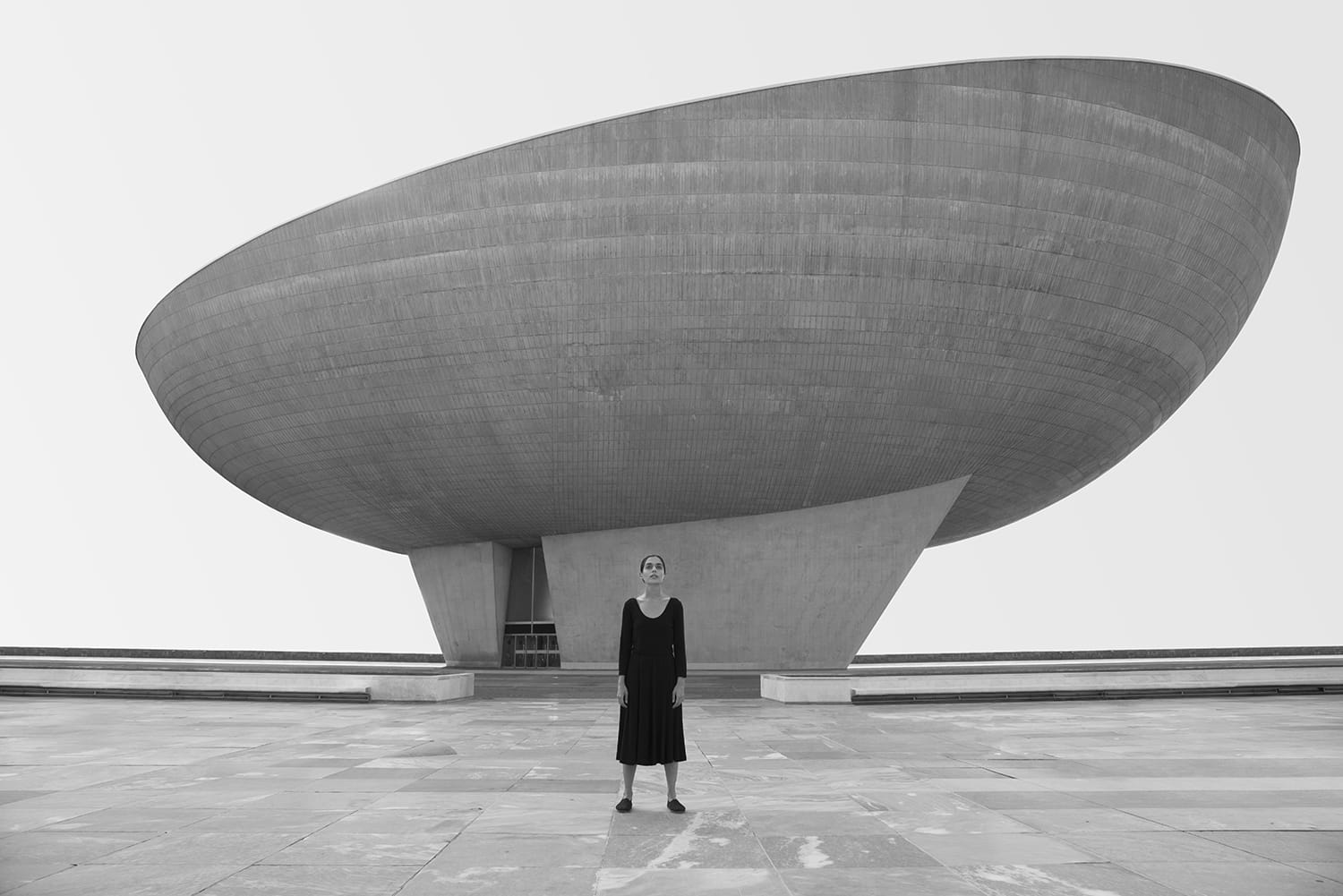 Person standing in front of a large modern, egg-shaped concrete building, wearing a black dress and shoes.
