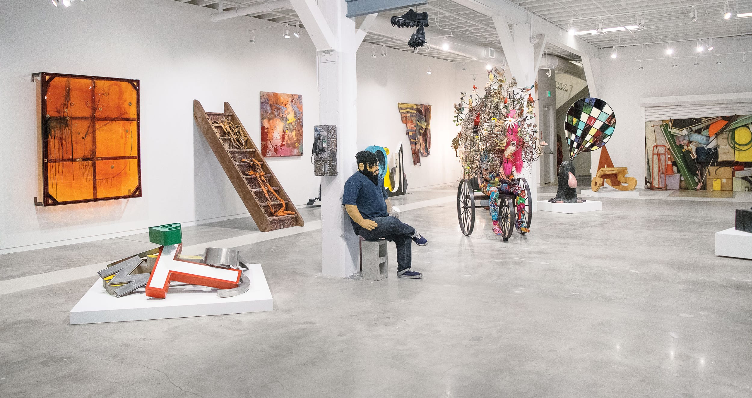 Contemporary art gallery with colorful sculptures, abstract paintings, and a person sitting on a bench in the center.