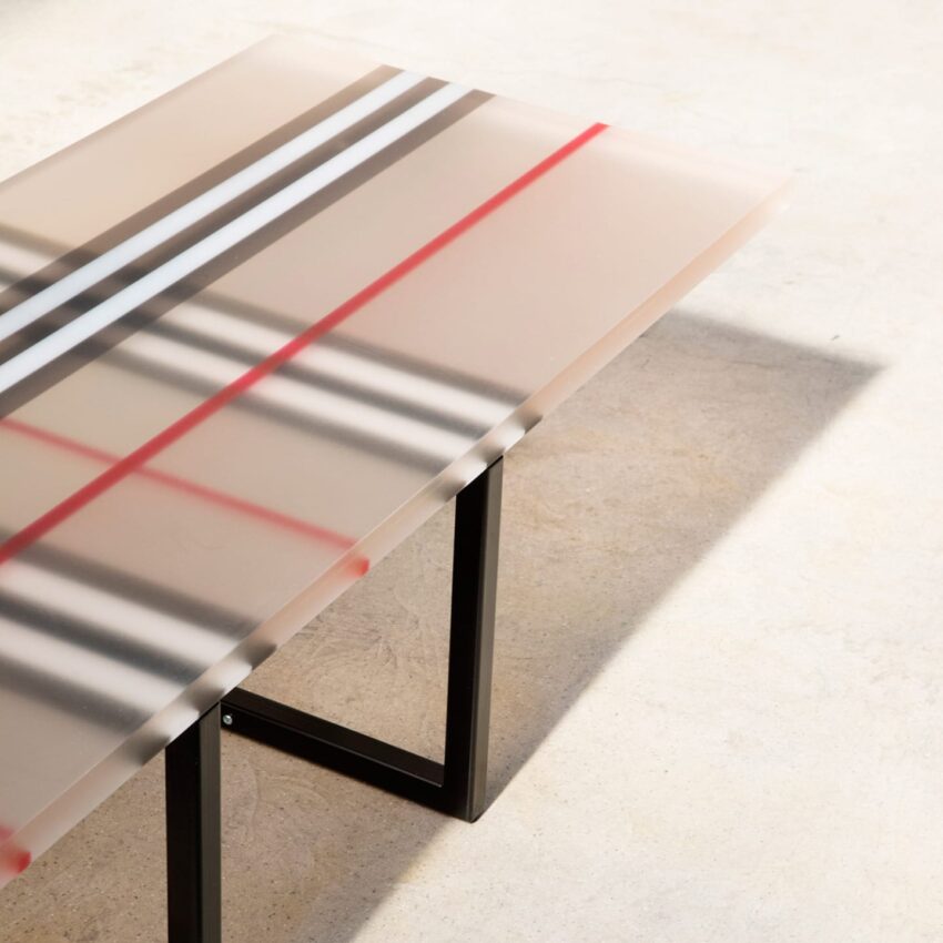 Modern glass table with black and red stripe pattern on metal legs, casting shadows on a light floor.