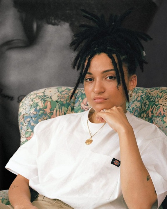 Person with dreadlocks sitting on a floral-patterned chair, wearing a white shirt and gold necklace, gazing at the camera.