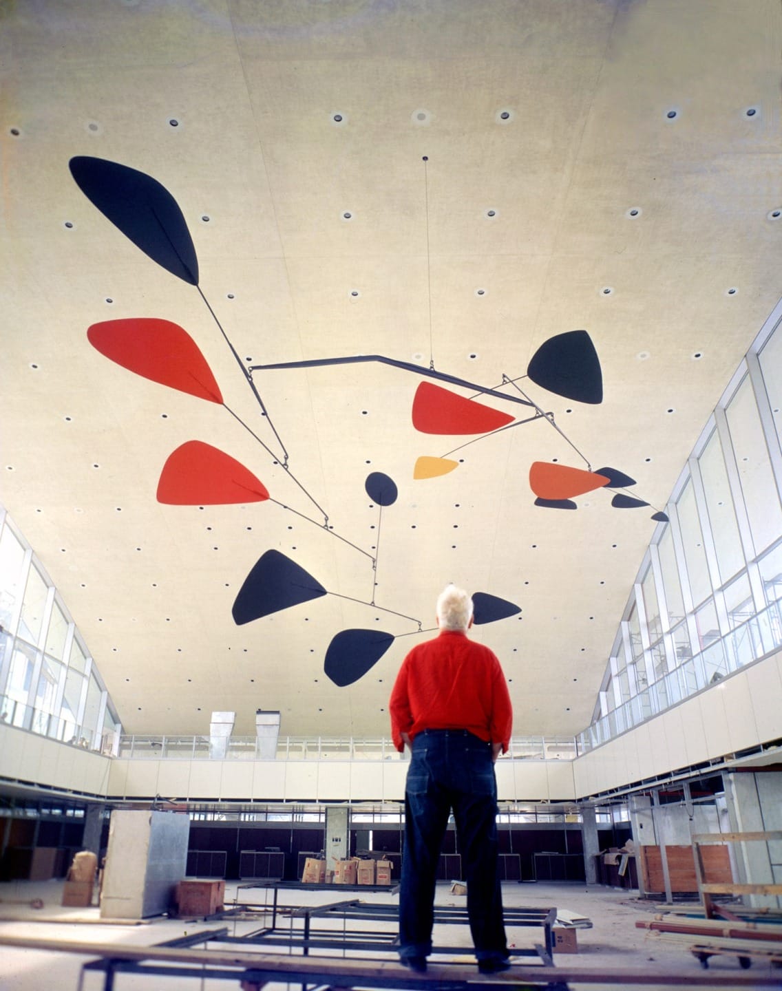 Man in red shirt standing beneath large abstract mobile sculpture in spacious, modern indoor setting with high ceiling.