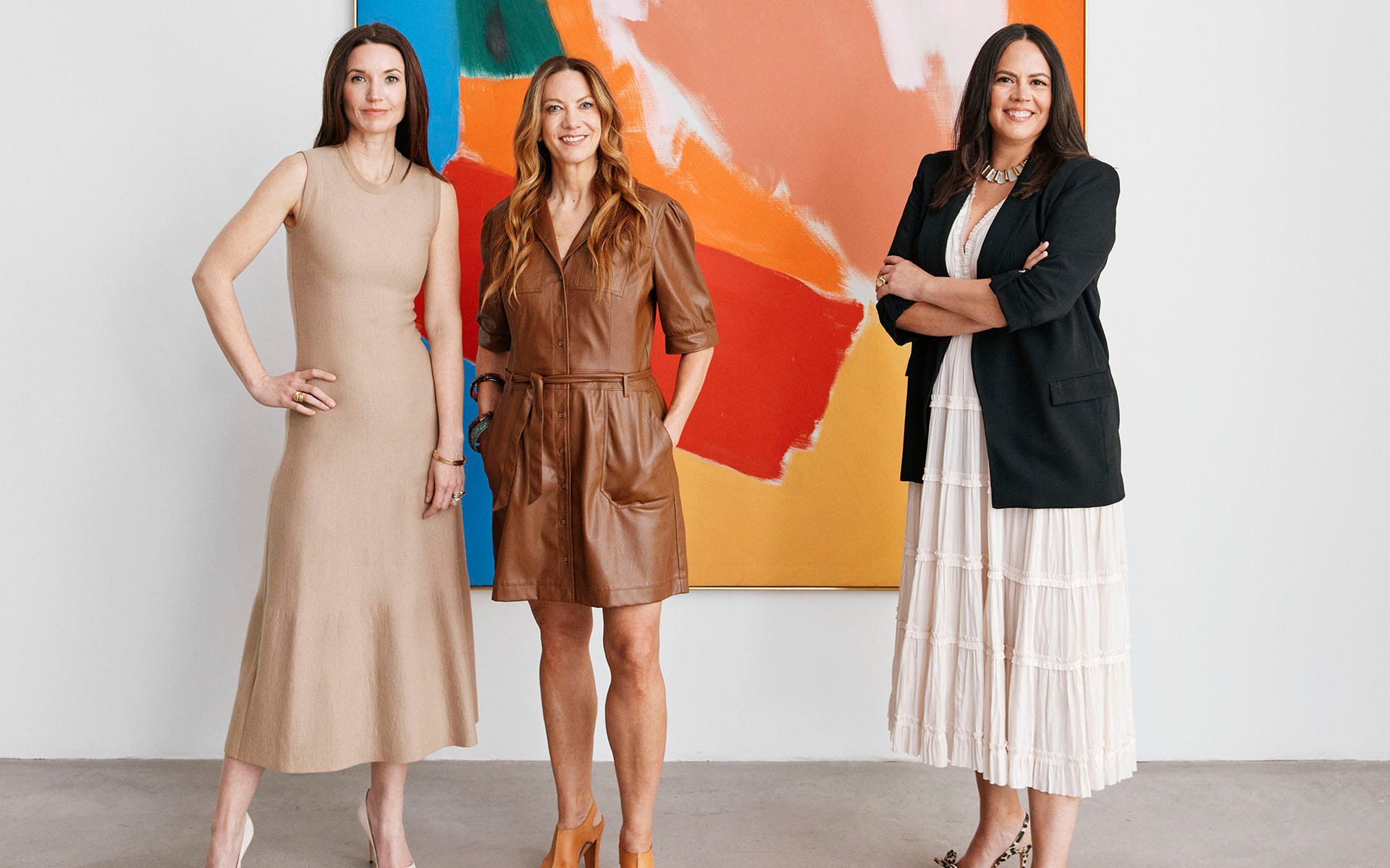 Three women standing confidently in front of a colorful abstract painting, each wearing stylish and elegant outfits.