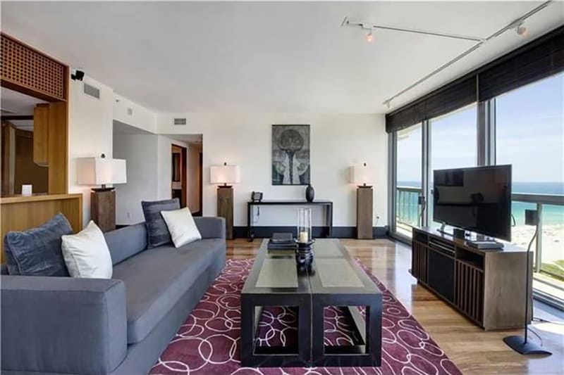 Modern living room with gray sofa, large windows, ocean view, flat-screen TV, contemporary decor, and red patterned rug.