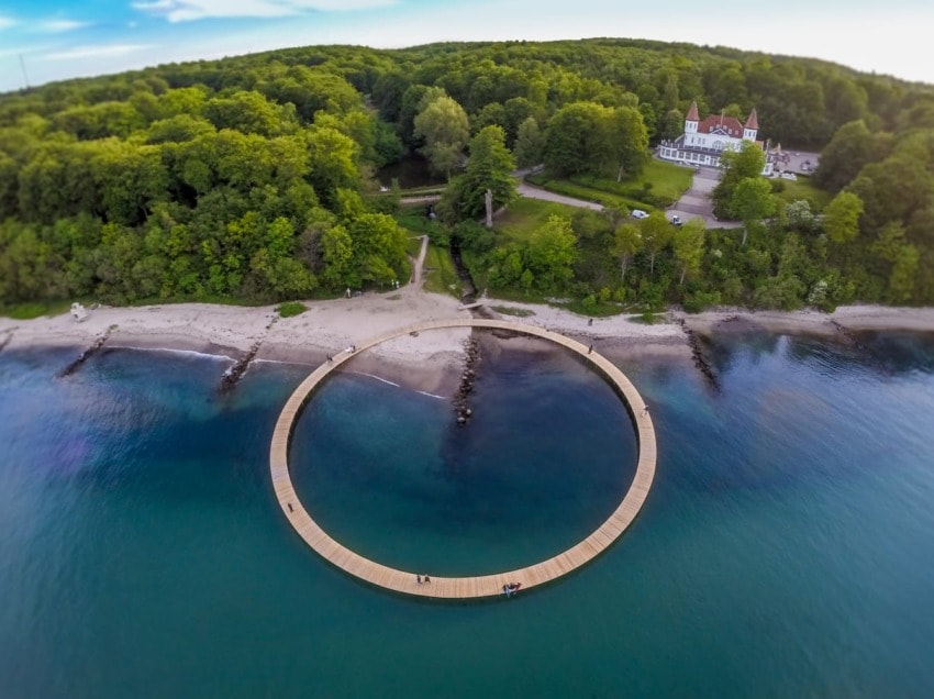 Aerial view of a circular wooden bridge over water near a forested shoreline with a building in the background