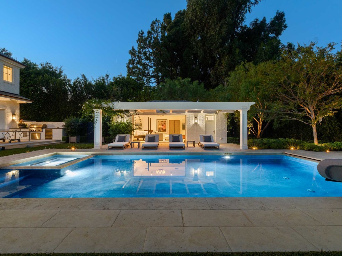 Luxury backyard pool with lounge chairs, pergola, and vibrant evening lighting surrounded by lush greenery.