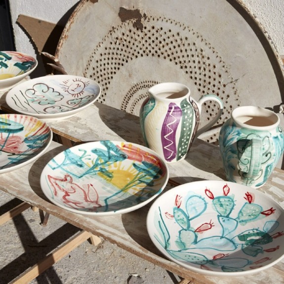 Hand-painted ceramic plates and jugs with colorful designs displayed on a wooden table outside.