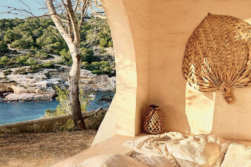 Seaside villa with woven wall art and pottery on the patio, overlooking a serene bay with lush greenery and rocky shoreline.