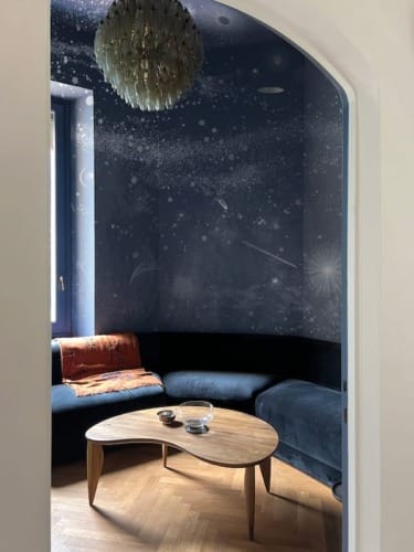 Cozy room with space-themed wallpaper, blue velvet couch, wooden coffee table, and modern chandelier.