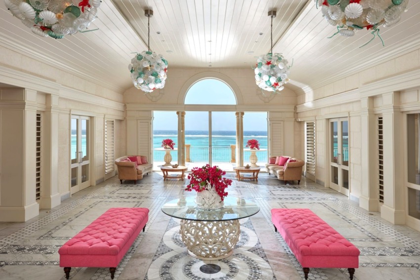 Luxurious beachside living room with pink seating, decorative chandeliers, and ocean view through large arched window.
