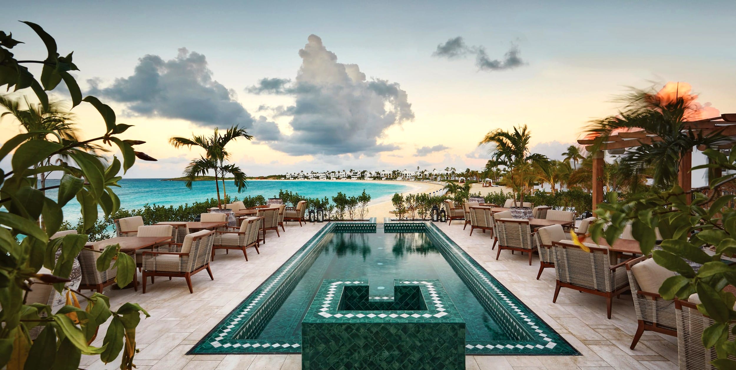 Oceanfront restaurant with infinity pool, palm trees, and seating area at sunset, overlooking a serene beach and cloudy sky.