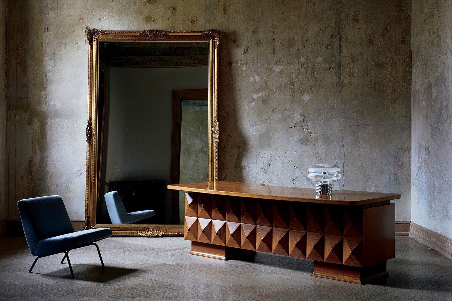 Vintage room with a large ornate mirror, geometric wooden desk, modern glass lamp, and a blue chair on wooden flooring.