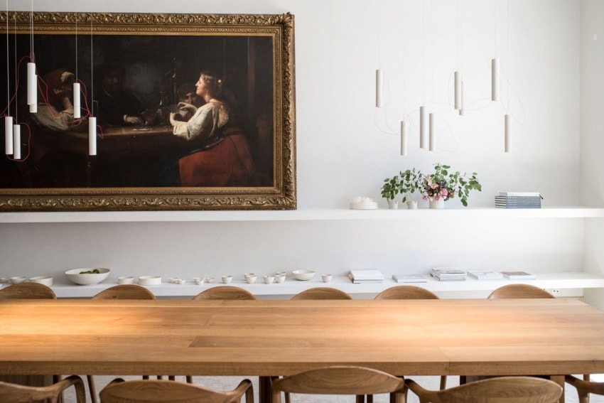 Modern dining area with wooden table, decorative hanging lights, framed classical painting, and shelf with plants and bowls.